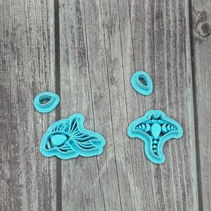 May include: A collection of light blue plastic cookie cutters in various shapes, including a fish, a manta ray, and two teardrop shapes. The cutters are arranged on a wooden surface with a natural wood grain.
