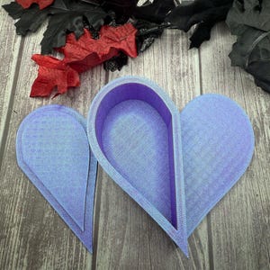 May include: Three lavender heart-shaped molds, possibly for crafting or baking, are displayed on a wooden surface. The molds have a textured appearance and a raised edge. Decorative black and red leaves are in the background.