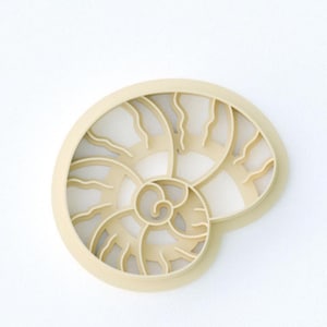 May include: A beige, nautilus-shaped wall decor piece. The design features a spiral pattern with curved lines and a circular outer edge. The piece is made of a solid material and is set against a white background.