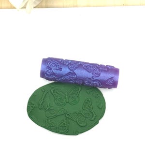 May include: A cylindrical purple rolling pin with a raised butterfly pattern and a flat, circular green piece of clay featuring an embossed butterfly design that matches the pattern on the rolling tool.