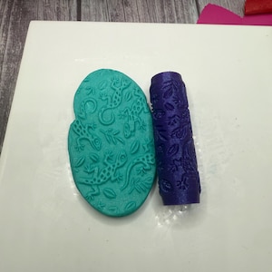May include: A teal, oval-shaped clay stamp with a lizard design and a purple rolling pin with a floral pattern. The tools are on a white surface. These tools are likely used for crafting or baking.