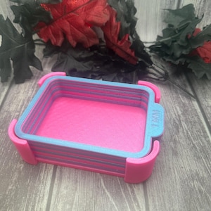 May include: A layered, rectangular tray in shades of pink, blue, and purple. The tray has a pink interior, blue edges, and purple layers. The text "1 MM" is visible on the side. The tray is likely used for organizing small items.