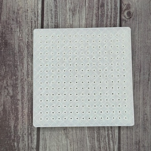 May include: A square, off-white plastic craft grid with numerous small, evenly spaced holes. The grid has a subtle diamond pattern and is set against a wooden background.