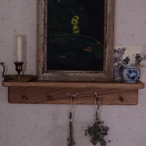 May include: A wooden shelf with three hooks, a framed painting of a green creature, a candle holder with a white candle, and a small blue and white ceramic vase with dried flowers.