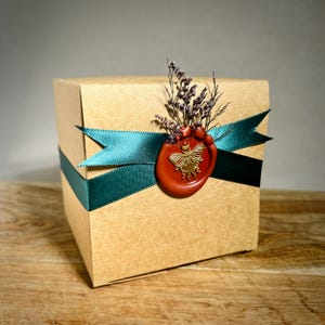 May include: A square, kraft paper gift box wrapped with a teal green ribbon. A red wax seal with a gold butterfly design and dried flowers adorn the box. The box is on a wooden surface.