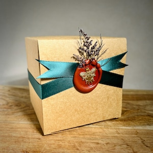 May include: A square cardboard gift box in a natural tan color, wrapped with a dark teal satin ribbon. A round red wax seal featuring a gold butterfly emblem secures the ribbon, accented with a small cluster of dried purple flowers on top.