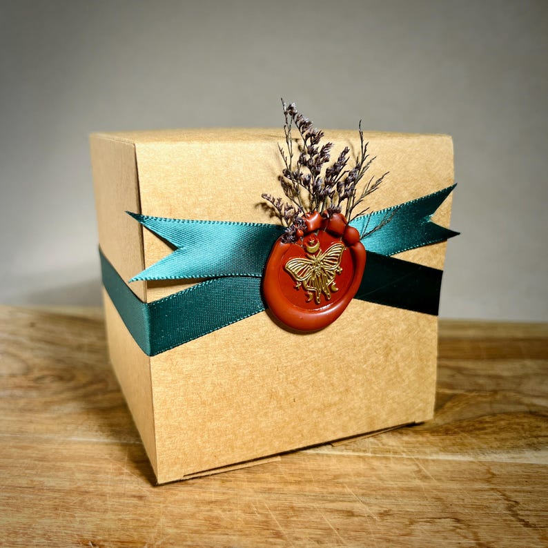 May include: A square cardboard gift box in a natural tan color, wrapped with a dark teal satin ribbon. A round red wax seal featuring a gold butterfly emblem secures the ribbon, accented with a small cluster of dried purple flowers on top.