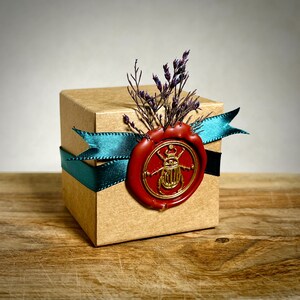 K&ouml;nnte beinhalten: Eine kleine, quadratische, braune Geschenkbox, verziert mit einem tealfarbenen Band und einem roten Wachssiegel mit einem goldenen K&auml;ferdesign. Die Box ist mit getrockneten lila Blumen verziert und schafft eine elegante und einzigartige Pr&auml;sentation.