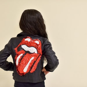 Puede incluir: Chaqueta negra de cuero sintético con un diseño de lengua y labios rojos y blancos con lentejuelas en la espalda. La chaqueta tiene un estilo motero clásico con cuello de solapa y cierre de cremallera. El modelo está de espaldas.