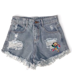 Puede incluir: Pantalones cortos vaqueros azul claro con detalles desgastados, incluyendo roturas y bordes deshilachados. Presentan un parche bordado colorido de un personaje parecido a un oso. Ropa casual.