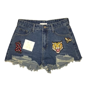 May include: Blue denim shorts with distressed edges and decorative patches. Patches include a red and black snake, a roaring tiger, and a bee. A white tag with text is attached to the shorts.