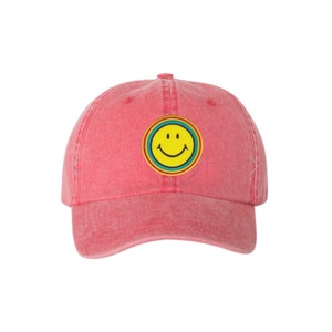 Puede incluir: Gorra de béisbol roja con visera curva. La gorra presenta un parche circular con una carita sonriente amarilla, delineada por anillos azules, verdes y rojos. El sombrero está hecho de un material suave y texturizado.