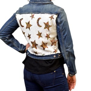 Può includere: Giacca di jeans blu con pannello posteriore in paillettes bianche con un motivo di stelle e lune crescenti dorate e marroni. La giacca ha maniche lunghe e un colletto classico. La modella indossa una maglietta nera e jeans blu.
