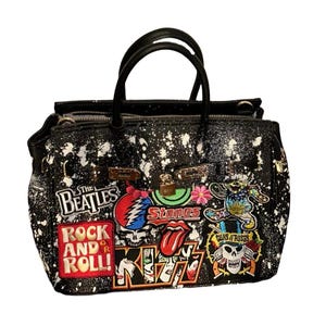 Op de afbeelding: Zwarte handtas met witte verfspatten en verschillende bandpatches, waaronder The Beatles, Grateful Dead, Rolling Stones, Guns N' Roses en KISS.