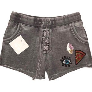 May include: Grey shorts with a lace-up front and two pockets. The shorts feature decorative patches of an ice cream cone, a pizza slice, and an eye. A blank tag is attached.