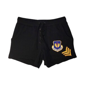 Può includere: Pantaloncini neri con vita con coulisse e dettaglio stringato sul davanti. I pantaloncini presentano una toppa con la scritta "US AIR FORCES IN EUROPE" e un design a galloni oro e giallo.