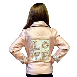 May include: A light pink faux leather jacket with the word "LOVE" spelled out on the back in colorful sequins. The jacket has a classic collar, long sleeves, and leopard print cuffs.