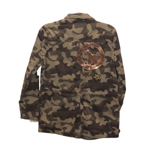 May include: Camouflage jacket with a peace sign and star patches. The jacket features a military-inspired design in shades of green, brown, and tan. The peace sign and star patches are embellished with sequins. The jacket has a button-up front and pockets.