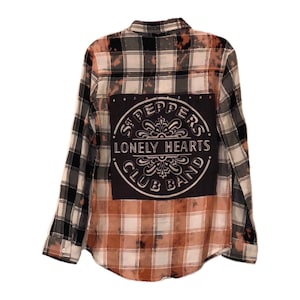 Może przedstawiać: Flanelowa koszula w czarno-białą kratę z rozpryskaną wybielaczem. Na plecach koszuli znajduje się czarna łatka z napisem "Sgt. Pepper's Lonely Hearts Club Band" w kolorze białym.