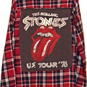 Puede incluir: Camisa de franela a cuadros rojos y negros con un parche gris en la espalda que presenta el logotipo de los Rolling Stones y el texto "U.S. Tour '78".