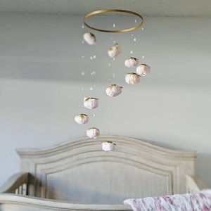 Puede incluir: Móvil para bebé con delicados adornos en forma de flor rosa claro suspendidos de un anillo dorado. El móvil cuelga sobre una cuna blanca con un cabecero decorativo. Pequeñas cuentas transparentes se intercalan entre las flores.