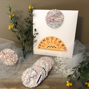 May include: A collection of map-themed coasters with scalloped edges. The coasters are arranged on a wooden surface with a white backdrop. The image also features a white card with the text "YOUNG'S FARM DESIGNS" and a floral accent.