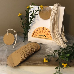 May include: A white paper bag with the text "YOUNG'S FARM DESIGNS" and a stack of scalloped-edge kraft paper tags. The bag has a cream-colored rope handle and is decorated with faux greenery and yellow flowers. A single tag hangs from a white string.