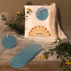 May include: A set of 15 light blue scalloped circle gift tags with a hole punched at the top. The tags are stacked on a wooden surface. A white twine tassel is included. The tags are perfect for adding a personal touch to gifts.