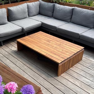 Puede incluir: Sofá seccional gris con mesa de centro de madera en una terraza de madera. El patio tiene un parterre con flores rosas y moradas. Una planta en maceta está al lado del sofá. La mesa de centro mide unos 100 cm de largo.