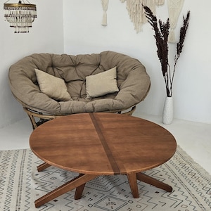 May include: A round wooden coffee table with a unique X-shaped base sits on a white and gray patterned rug in a living room. A brown, round,  oversized chair with two pillows is in the background, along with a wicker chair.