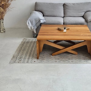 Puede incluir: Una mesa de centro de madera con una base en forma de X única. La mesa tiene un acabado marrón claro y está sentada sobre una alfombra estampada frente a un sofá gris.