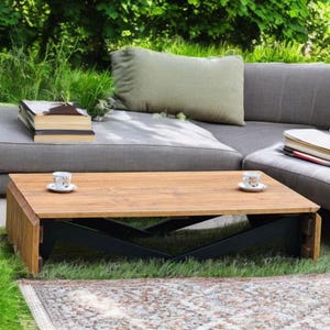 Puede incluir: Una mesa de centro rectangular de madera con acabado natural y soportes de metal negro. Dos tazas y platillos pequeños están sobre la mesa. Un sofá seccional gris y hierba verde en el fondo.