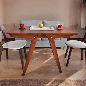 Könnte beinhalten: Ein Esstisch aus Holz mit einer hellen Holzoptik und einer weißen Tischplatte. Der Tisch hat ein Klappblatt-Design und ist mit zwei Stühlen ausgestattet. Die Stühle haben dunkle Holzrahmen und weiße Sitze.