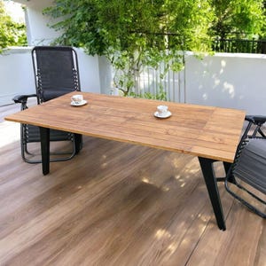 May include: A wooden outdoor dining table with black legs and two matching chairs. Two small teacups and saucers sit on the table. The table is made of light brown wood and is surrounded by greenery.
