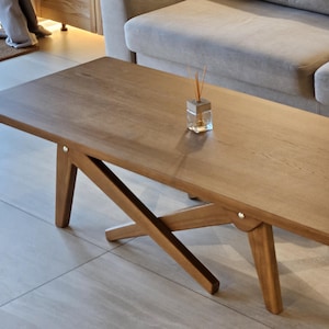 May include: A rectangular wooden coffee table with a light brown finish. The table has a unique X-shaped base made of four wooden legs. The table is sitting in front of a grey sofa.