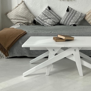May include: White folding coffee table with a simple, modern design. The table has a rectangular top and a unique X-shaped base. The table is sitting on a white floor in front of a couch with pillows.