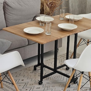 May include: A wooden table with a black metal frame and four white chairs. The table is set with four white plates and four clear glasses. The table is in a room with a gray couch and a white rug.