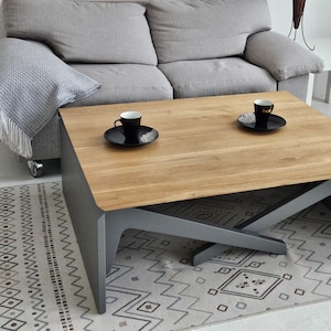 May include: A modern coffee table with a light wood top and grey legs. The table has a unique design with the legs crossing over each other. Two black and white coffee cups are on the table.