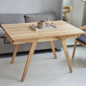 Può includere: Un tavolo in legno chiaro con un piano rettangolare e gambe inclinate. Il tavolo ha un vassoio sopra con un piccolo vaso in ceramica e un barattolo di vetro. Due sedie con telai in legno chiaro e cuscini grigio scuro sono accanto al tavolo.