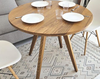 Könnte beinhalten: Ein runder Holztisch mit vier Beinen und einem weißen gemusterten Teppich darunter. Auf dem Tisch stehen vier weiße Teller und vier Gläser, sowie ein weißer Stuhl.