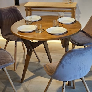 May include: A round wooden table with four chairs. The table has a light brown finish and the chairs are upholstered in a light gray fabric. There are four white plates and glasses on the table.