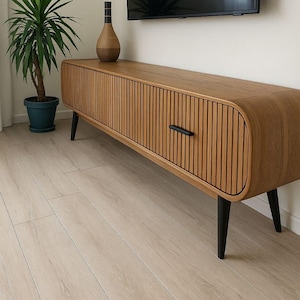 May include: A wooden TV cabinet in a mid-century modern style, featuring a ribbed front and black legs. A decorative vase sits on top. A large flat-screen TV is mounted on the wall above the cabinet. A potted plant is to the left.