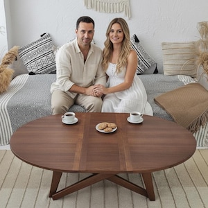 May include: A dark brown, oval-shaped coffee table with a cross-legged base. The table is set with two cups of coffee and a plate of cookies. A couple is seated behind the table on a sofa with decorative pillows.