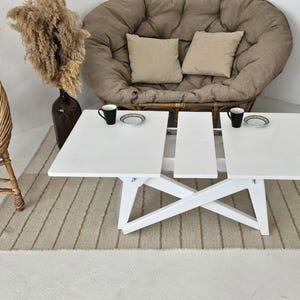 May include: White extendable coffee table with a white, woven rug underneath. The table has a white, X-shaped base and two white rectangular tabletops. The table is in a living room setting with a brown, round chair and a white chair in the background.