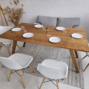 Könnte beinhalten: Ein Esstisch aus Holz mit einem weißen gemusterten Teppich darunter. Der Tisch ist mit weißen Tellern, Besteck und Gläsern gedeckt. Es gibt vier weiße Stühle um den Tisch herum und ein graues Sofa im Hintergrund.