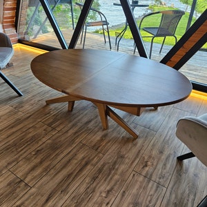Könnte beinhalten: Ein ovaler Couchtisch aus Holz mit einem einzigartigen X-förmigen Gestell. Der Tisch ist aus dunklem Holz gefertigt und hat eine glatte, polierte Oberfläche. Der Tisch steht auf einem Holzboden.
