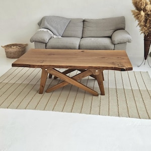 May include: A wooden coffee table with a natural finish and a live edge design. The table has a cross-legged base and sits on a striped rug. A grey sofa and woven basket are in the background.