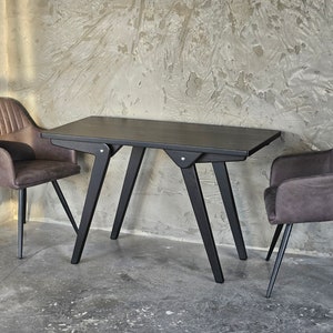 Puede incluir: Una mesa de madera negra con una tapa rectangular y un diseño de patas angulares único. Dos sillas tapizadas en marrón con patas de metal negro se colocan a cada lado de la mesa.