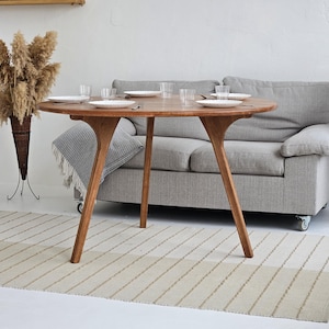 Pode incluir: Uma mesa de jantar redonda de madeira com acabamento natural, com pratos brancos e copos transparentes. A mesa tem um design &uacute;nico de tr&ecirc;s pernas. Um sof&aacute; cinza e um tapete listrado est&atilde;o no fundo. Plantas secas decorativas nos cantos.