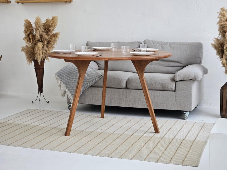 Pode incluir: Uma mesa de jantar redonda de madeira com acabamento natural, com pratos brancos e copos transparentes. A mesa tem um design &uacute;nico de tr&ecirc;s pernas. Um sof&aacute; cinza e um tapete listrado est&atilde;o no fundo. Plantas secas decorativas nos cantos.